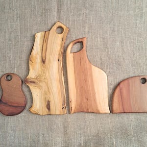 Puede incluir: Cuatro tablas de cortar de madera con bordes naturales y orificios de agarre integrados. Las tablas var&iacute;an en forma y tama&ntilde;o, presentando patrones de vetas de madera marr&oacute;n claro y oscuro, desde una tabla peque&ntilde;a redondeada hasta una losa alta e irregular.