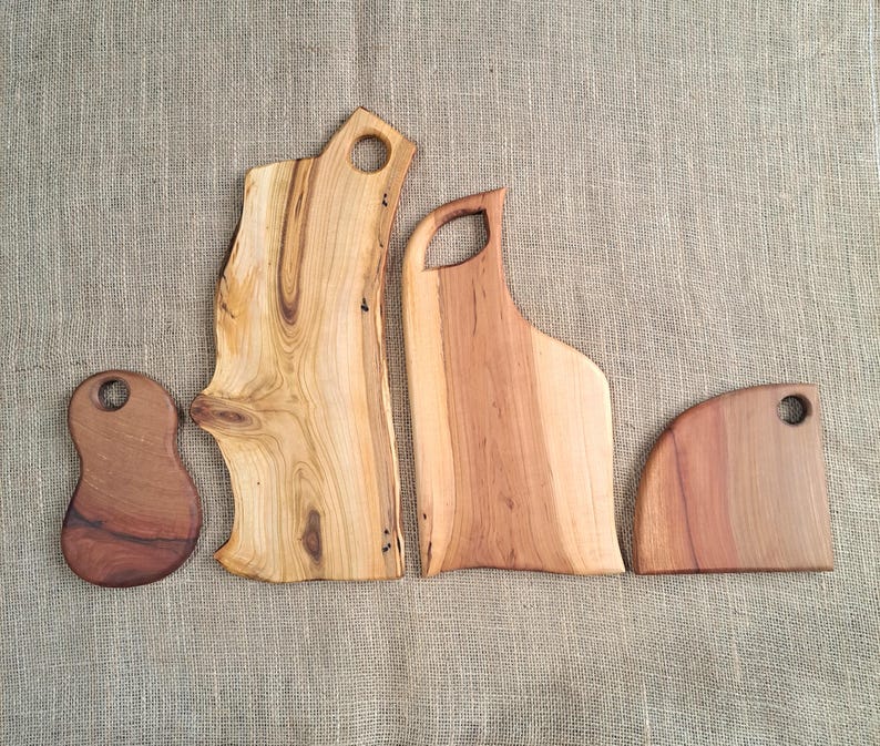 Puede incluir: Cuatro tablas de cortar de madera con bordes naturales y orificios de agarre integrados. Las tablas var&iacute;an en forma y tama&ntilde;o, presentando patrones de vetas de madera marr&oacute;n claro y oscuro, desde una tabla peque&ntilde;a redondeada hasta una losa alta e irregular.