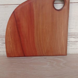 Puede incluir: Una tabla de cortar de madera en forma de cuarto de c&iacute;rculo con un patr&oacute;n de veta suave de color marr&oacute;n rojizo. La tabla presenta un recorte circular cerca de la esquina superior derecha para colgarla, con un borde inferior plano.