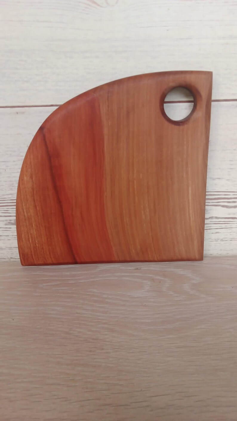 Puede incluir: Una tabla de cortar de madera en forma de cuarto de c&iacute;rculo con un patr&oacute;n de veta suave de color marr&oacute;n rojizo. La tabla presenta un recorte circular cerca de la esquina superior derecha para colgarla, con un borde inferior plano.