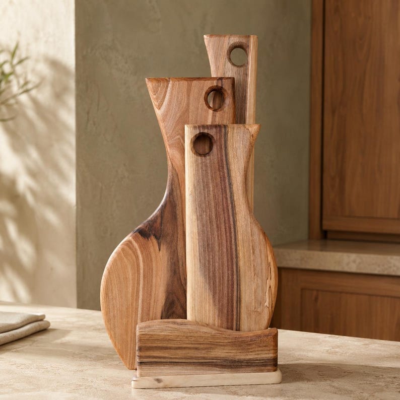 Op de afbeelding: Een set van drie verticale houten snijplanken met natuurlijke houtnerven, elk voorzien van een rond gat aan de bovenkant om op te hangen. De planken staan in een getrapte opstelling in een rechthoekige houten standaard.