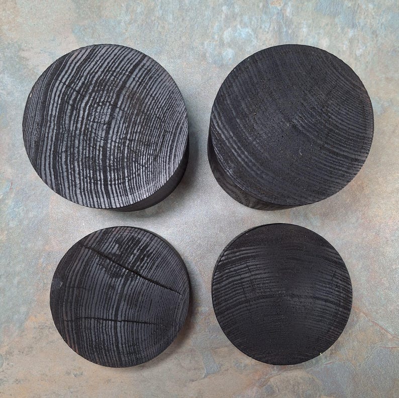 May include: Four circular, black wooden coasters with visible wood grain. Two coasters display a lighter, more prominent grain, while the other two have a darker, less defined grain. The coasters are arranged in a square formation.