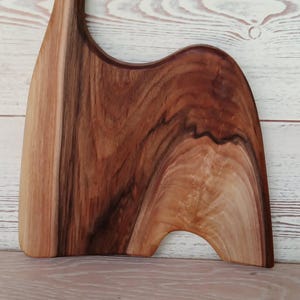 May include: A wooden serving board with a natural grain pattern, featuring a tall, curved handle with a circular cutout at the top. The board has an asymmetrical, organic shape with a smooth, polished finish showing light and dark brown wood tones.