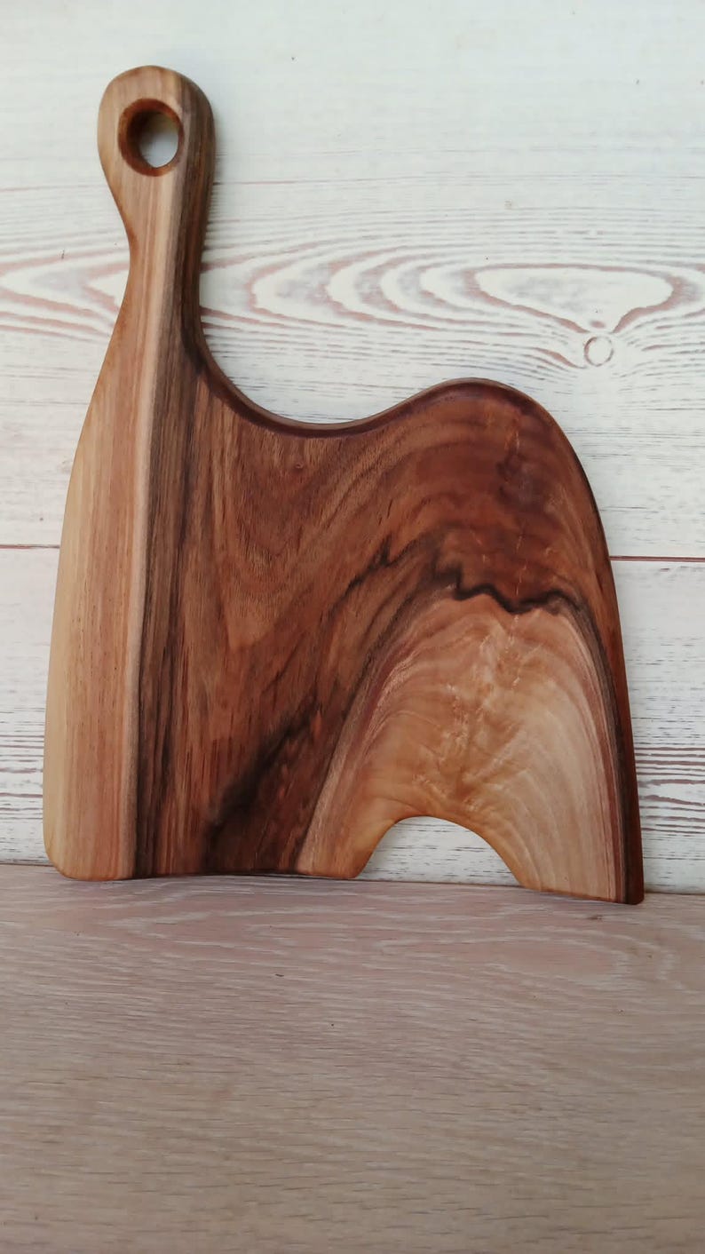 May include: A wooden serving board with a natural grain pattern, featuring a tall, curved handle with a circular cutout at the top. The board has an asymmetrical, organic shape with a smooth, polished finish showing light and dark brown wood tones.
