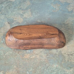 May include: A handcrafted, wooden serving tray with a natural, organic shape. The tray is made of dark brown wood with visible grain patterns and a smooth, polished finish. The tray is approximately 12 inches long and 6 inches wide.