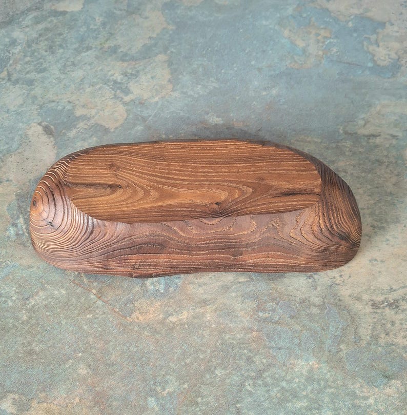 May include: A handcrafted, wooden serving tray with a natural, organic shape. The tray is made of dark brown wood with visible grain patterns and a smooth, polished finish. The tray is approximately 12 inches long and 6 inches wide.