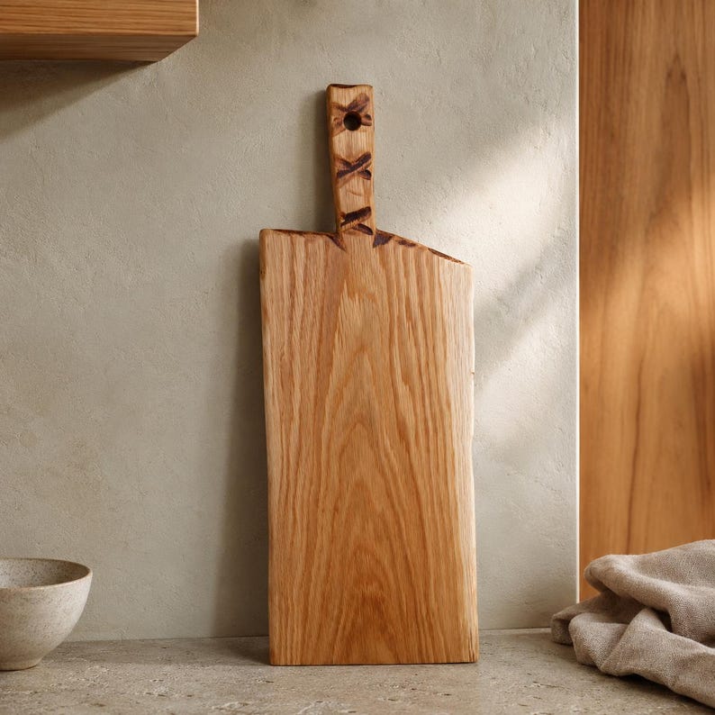 Pode incluir: Uma t&aacute;bua de corte retangular de madeira com um padr&atilde;o de gr&atilde;o de madeira castanho-claro natural. A t&aacute;bua apresenta uma pega vertical no topo com um orif&iacute;cio circular e detalhes decorativos em ponto cruz castanho-escuro na pega.