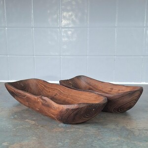 May include: Two handcrafted wooden bowls with a natural, organic shape. The bowls are made of dark brown wood, showcasing the grain and texture. They are placed side by side, suggesting they are a set. The bowls have a rustic, earthy aesthetic.