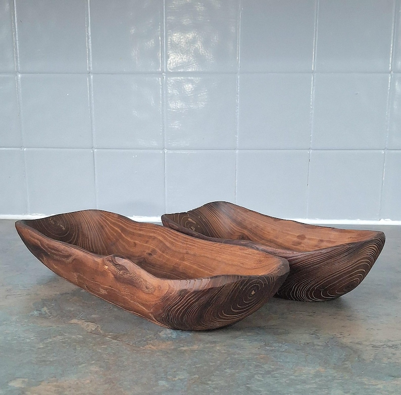 May include: Two handcrafted wooden bowls with a natural, organic shape. The bowls are made of dark brown wood, showcasing the grain and texture. They are placed side by side, suggesting they are a set. The bowls have a rustic, earthy aesthetic.