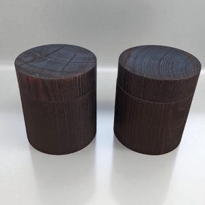 May include: Two dark brown wooden boxes with lids. The boxes are cylindrical with a textured wood grain pattern. The lids have a circular grain pattern. The boxes are displayed on a white surface.