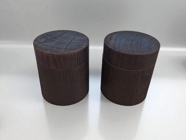 May include: Two dark brown wooden boxes with lids. The boxes are cylindrical with a textured wood grain pattern. The lids have a circular grain pattern. The boxes are displayed on a white surface.