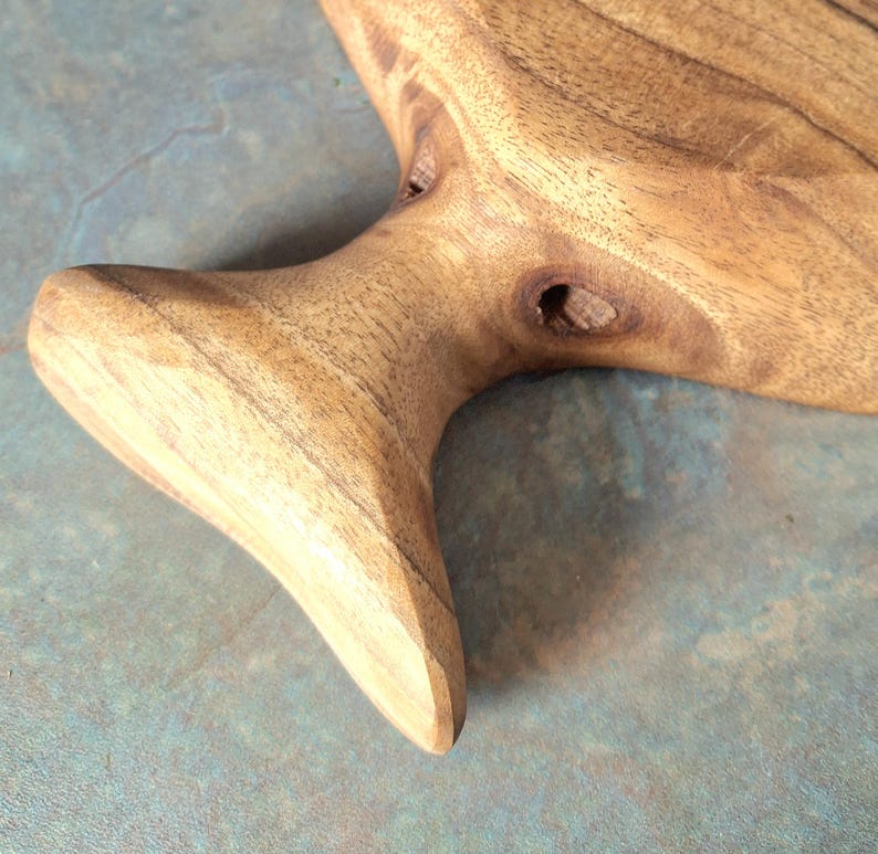 May include: Close-up of a handcrafted wooden serving board with a natural, organic shape. The board features a light brown colour with visible wood grain and a handle-like extension. The surface is smooth, showcasing the natural beauty of the wood.