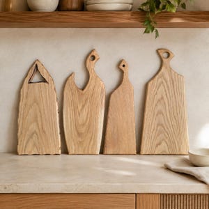 Può includere: Quattro taglieri in legno chiaro con forme distinte e venature visibili. I taglieri presentano sagome varie, tra cui uno con un intaglio triangolare, due con fori per l'impugnatura e uno con un design dal bordo liscio e affusolato.