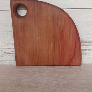 Puede incluir: Una tabla de cortar de madera en forma de cuarto de c&iacute;rculo con una veta suave de color marr&oacute;n rojizo y un orificio circular cerca de la esquina superior izquierda para colgarla. La tabla presenta un borde vertical recto, una base horizontal y un borde exterior curvo.