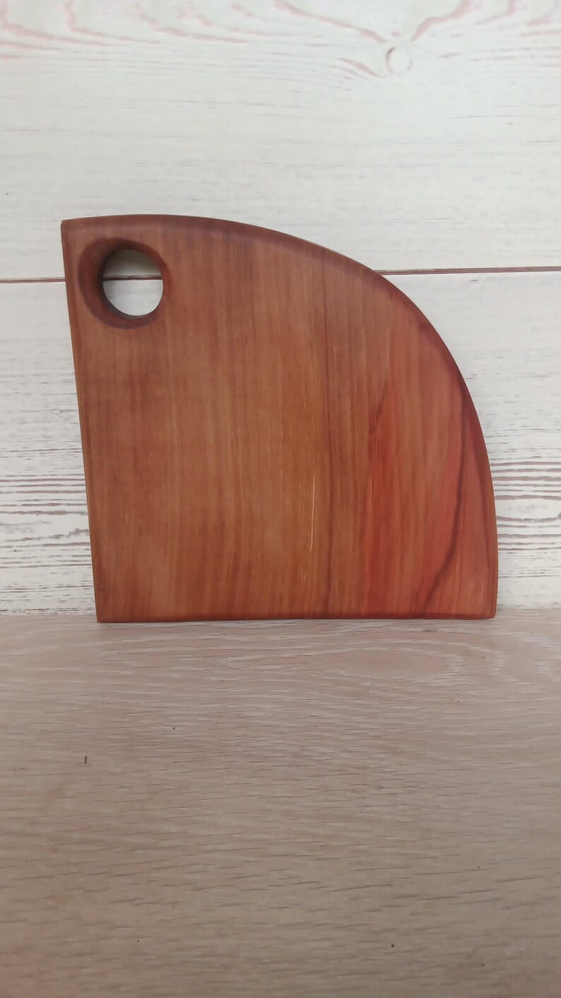 Puede incluir: Una tabla de cortar de madera en forma de cuarto de c&iacute;rculo con una veta suave de color marr&oacute;n rojizo y un orificio circular cerca de la esquina superior izquierda para colgarla. La tabla presenta un borde vertical recto, una base horizontal y un borde exterior curvo.