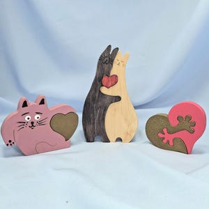 以下が含まれることがあります： 木製の猫をテーマにした装飾品3点。ハート型の切り抜きがあるピンク色の猫の形をしたアイテム1点。抱き合っている2匹の猫のフィギュア、濃い茶色と薄い茶色、赤いハート付き。金色のデザインのピンク色のハート型のアイテム。