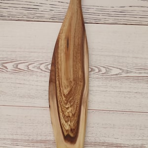 May include: A long, narrow wooden serving board with a tapered handle featuring a circular hole at the top. The board displays a prominent natural wood grain pattern with contrasting light and dark brown tones, showcasing a smooth, polished finish.