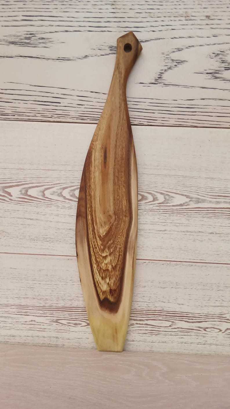 May include: A long, narrow wooden serving board with a tapered handle featuring a circular hole at the top. The board displays a prominent natural wood grain pattern with contrasting light and dark brown tones, showcasing a smooth, polished finish.