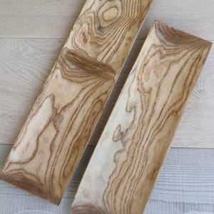 May include: Two rectangular wooden trays with a light tan base color and prominent dark brown wood grain patterns. The trays have a smooth, elongated shape with slightly curved edges, showcasing the natural texture and organic lines of the wood.