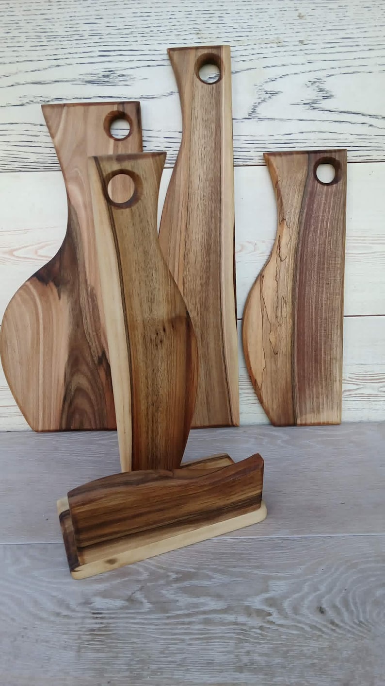 Op de afbeelding: Een set van vier houten serveerplanken met natuurlijke houtnerven en ronde handvatopeningen, vergezeld door een rechthoekige houten standaard. De planken hebben vari&euml;rende vormen en bruintinten en staan rechtop in een groep.