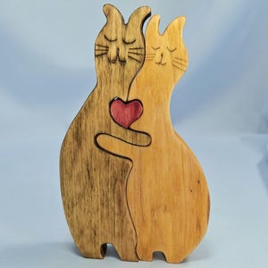 以下が含まれることがあります： 抱き合う2匹の猫の木製置物。片方の猫は濃い茶色、もう片方は明るい木の色合い。赤いハートが間に埋め込まれ、愛を象徴しています。愛らしい装飾品です。