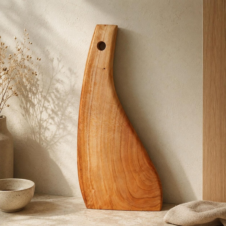 May include: A tall, light brown wooden cutting board with a curved, asymmetrical silhouette and a circular hole near the top for hanging. The board features a smooth, polished surface with visible natural wood grain patterns throughout the piece.
