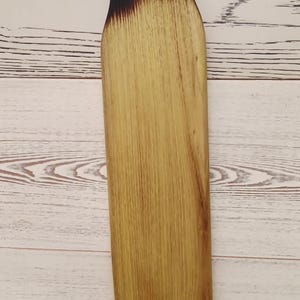 Pode incluir: Uma t&aacute;bua de servir de madeira longa e estreita com um acabamento de gr&atilde;o de madeira castanho claro natural. O topo apresenta um orif&iacute;cio circular para a pega, com a madeira &agrave; volta da pega e a sec&ccedil;&atilde;o superior a transitar para um preto carbonizado.