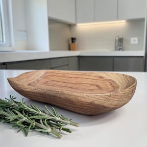 May include: An oval wooden bowl with a natural wood grain finish. The bowl is empty and rests on a white countertop, with a sprig of rosemary in front. The kitchen setting suggests a culinary theme.