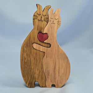 以下が含まれることがあります： 抱き合う2匹の猫の木製置物。猫は異なる木の色合いで作られ、中央に赤いハートがあります。愛らしい手作りの装飾品です。