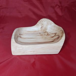 May include: A wooden bowl with a natural finish. The bowl has an irregular shape with a raised section. The wood grain is visible, adding to its rustic charm. The bowl is set against a red background. The bowl's dimensions are approximately 30 cm in length.