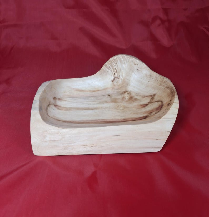 May include: A wooden bowl with a natural finish. The bowl has an irregular shape with a raised section. The wood grain is visible, adding to its rustic charm. The bowl is set against a red background. The bowl's dimensions are approximately 30 cm in length.