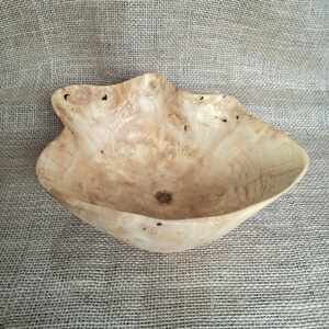 Op de afbeelding: Een handgemaakte houten kom met een natuurlijke, lichtgekleurde afwerking. De kom heeft een organische, onregelmatige vorm met een textuur en zichtbare houtnerf. De kom wordt waarschijnlijk gebruikt voor decoratieve of serveerdoeleinden.