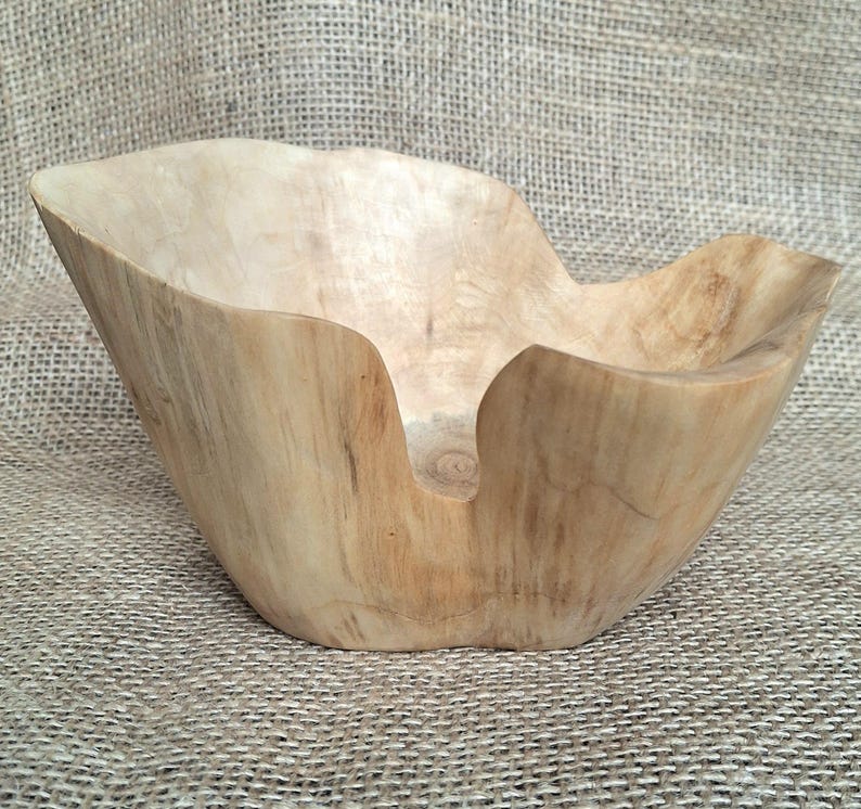 Op de afbeelding: Een handgesneden houten kom met een natuurlijke, lichtbruine afwerking. De kom heeft een organische, asymmetrische vorm met een brede opening en een gladde binnenkant. De houtnerf is zichtbaar.