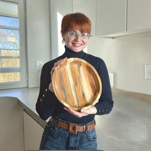 Può includere: Un vassoio rotondo in legno con un motivo a venature naturali. Il vassoio è tenuto da una persona che indossa un dolcevita nero e jeans blu. Il vassoio ha un diametro di circa 30 cm.