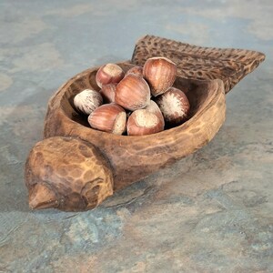 Pu&ograve; includere: Una ciotola di legno intagliato a forma di uccello, piena di nocciole. La ciotola ha una finitura naturale e rustica con venature del legno visibili e una coda intagliata. Le nocciole sono marroni con macchie chiare.