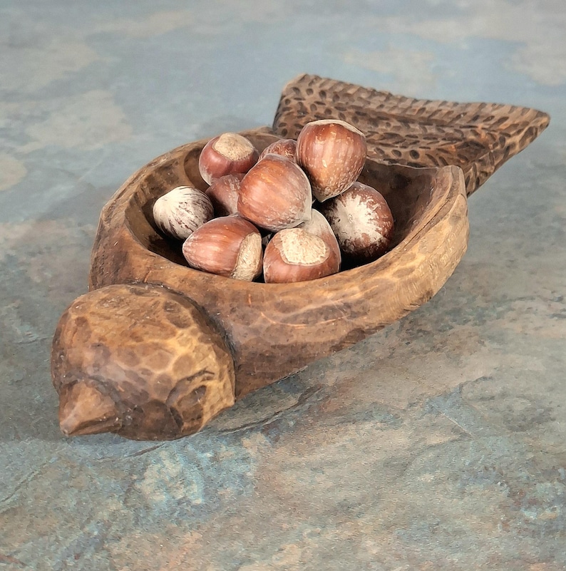 Pu&ograve; includere: Una ciotola di legno intagliato a forma di uccello, piena di nocciole. La ciotola ha una finitura naturale e rustica con venature del legno visibili e una coda intagliata. Le nocciole sono marroni con macchie chiare.