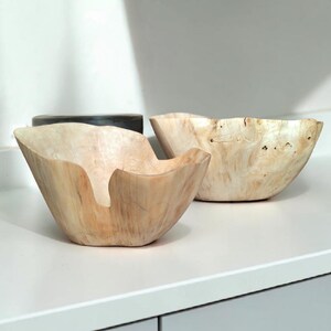 Op de afbeelding: Twee houten kommen met natuurlijke houtnerf. De kommen hebben een unieke, organische vorm, waarbij &eacute;&eacute;n kom een opvallende uitsparing heeft. De kommen zijn lichtbruin van kleur en lijken handgemaakt.