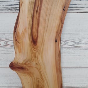 Puede incluir: Una tabla de servir de madera de color marr&oacute;n claro con un borde natural y un nudo prominente en el lado izquierdo. La tabla presenta una superficie lisa y pulida con patrones de grano de madera visibles y un asa circular recortada.