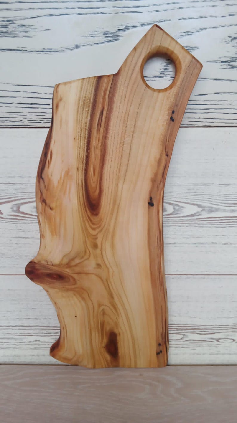 Puede incluir: Una tabla de servir de madera de color marr&oacute;n claro con un borde natural y un nudo prominente en el lado izquierdo. La tabla presenta una superficie lisa y pulida con patrones de grano de madera visibles y un asa circular recortada.