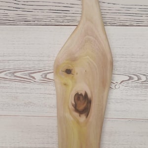 Pode incluir: Uma t&aacute;bua de servir de madeira clara com borda natural, apresentando um n&oacute; de madeira escura proeminente no centro e um n&oacute; menor acima. A t&aacute;bua tem um formato alongado e assim&eacute;trico com uma al&ccedil;a no topo contendo um orif&iacute;cio circular.