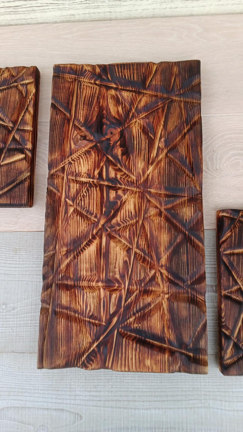 May include: Three rectangular wooden wall art pieces with a geometric, carved design. The wood has a dark brown, burnt finish, with visible wood grain. The carved lines create a textured, dimensional effect, adding visual interest to the decor.