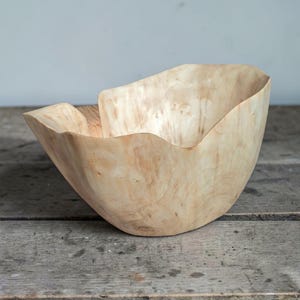 Op de afbeelding: Een handgemaakte houten kom met een natuurlijke, lichtgeleurde afwerking. De kom heeft een organische, asymmetrische vorm met een golvende rand. De houtnerf is zichtbaar, wat bijdraagt aan de rustieke uitstraling. De kom is waarschijnlijk voor decoratieve of serveerdoeleinden.