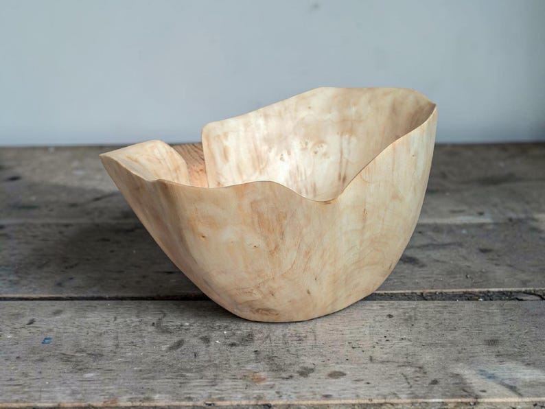 Op de afbeelding: Een handgemaakte houten kom met een natuurlijke, lichtgeleurde afwerking. De kom heeft een organische, asymmetrische vorm met een golvende rand. De houtnerf is zichtbaar, wat bijdraagt aan de rustieke uitstraling. De kom is waarschijnlijk voor decoratieve of serveerdoeleinden.
