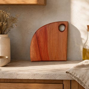 Puede incluir: Una tabla de cortar de madera en forma de cuarto de c&iacute;rculo con una veta suave de color marr&oacute;n rojizo y un orificio circular cerca de la esquina superior derecha para colgarla. La tabla presenta una base plana y un borde superior curvo.