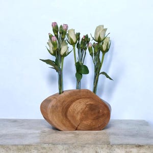 Puede incluir: Un jarrón de madera con tres tubos de vidrio para sostener flores. El jarrón está hecho de una rebanada de tronco de árbol y tiene un acabado de madera natural. Rosas blancas con centros rosados ​​están dispuestas en el jarrón.