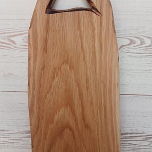 Pode incluir: Uma t&aacute;bua de servir de madeira castanho-clara com uma borda natural e uma pega triangular recortada no topo. A t&aacute;bua apresenta um padr&atilde;o de gr&atilde;o de madeira proeminente e uma superf&iacute;cie lisa e polida, real&ccedil;ando a textura org&acirc;nica da madeira.