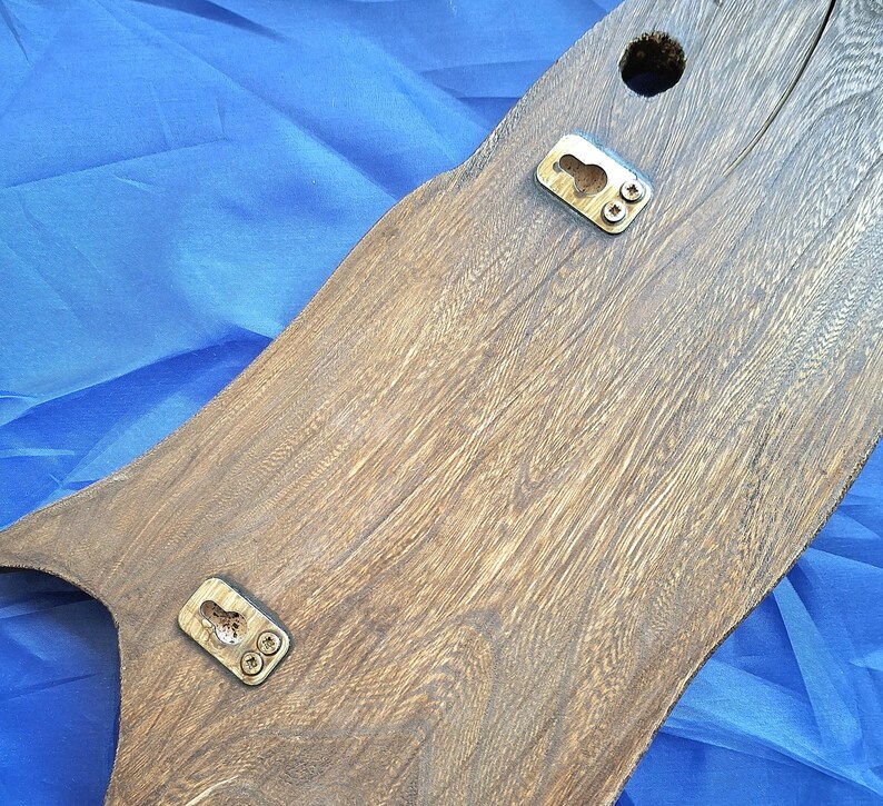 May include: A wooden wall hanging with a natural wood grain finish. The piece has a unique, organic shape and features two metal keyhole hangers and a drilled hole for mounting. The wood is a warm brown color.