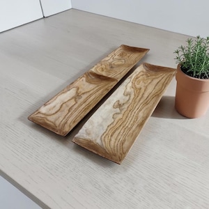 May include: Two rectangular wooden serving trays with a distinct light and dark brown wood grain pattern, positioned side by side. Next to the trays is a small, light orange terracotta pot containing a green plant with thin, needle-like leaves.