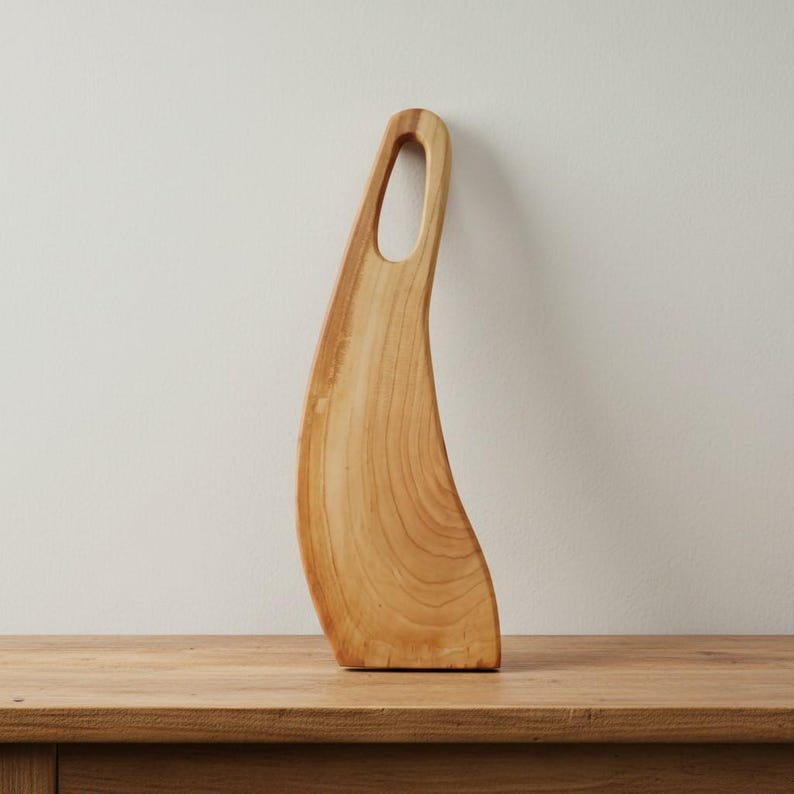 May include: A light brown wooden serving board with a curved, elongated shape and a smooth finish. The board features a distinct natural wood grain pattern and an integrated handle with an oval cutout at the top for easy gripping and hanging.