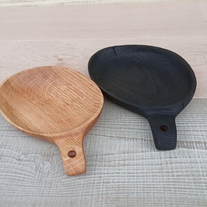 K&ouml;nnte beinhalten: Zwei Holzserviertabletts mit Griffen. Eines ist naturfarben, das andere schwarz. Beide haben eine runde Schalenform und einen Griff mit einem Loch zum Aufh&auml;ngen. Die Tabletts liegen auf einer hellen Holzoberfl&auml;che.